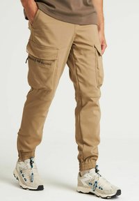 Beige cargobroek met meerdere zakken, elastische boorden en een duurzame stof. Geweldig te combineren met lichtgrijze sneakers met blauwe accenten.