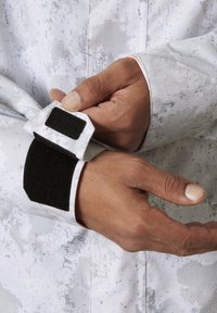 Bílá bunda s texturovaným, tečkovaným vzorem. Obsahuje nastavitelné suché zipy na zápěstí s černým polstrováním. Detailní záběr na ruku, která zajišťuje zápěstí.
