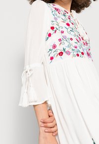 Vestido blanco con bordado floral en rojo, rosa y verde. Presenta detalles de lazo en los extremos de las mangas y un diseño plisado para añadir textura.