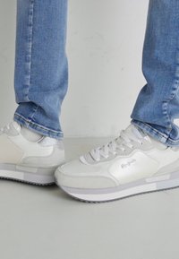 White sneakers with mixed material design, featuring glossy accents and textured overlays. Paired with light blue denim jeans.