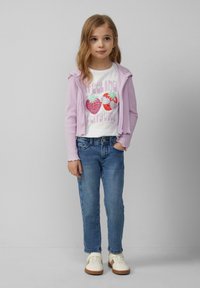 Paarse geribde hoodie met rits over een witte T-shirt met een aardbeienafbeelding, gecombineerd met blauwe denim en crèmekleurige sneakers met bruine zolen.