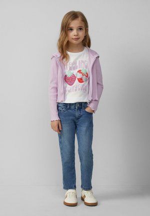 Paarse geribde hoodie met rits over een witte T-shirt met een aardbeienafbeelding, gecombineerd met blauwe denim en crèmekleurige sneakers met bruine zolen.