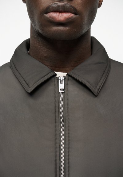 Gros plan sur le bas du visage et le cou d'une personne portant une veste gris foncé zippée avec un col pointu, par-dessus une chemise claire.