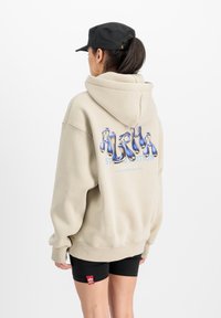 Beige oversized hoodie met een grafisch ontwerp "ALPHA" in blauw en zilver op de achterkant. Gecombineerd met zwarte shorts. Zwarte pet achterstevoren gedragen.