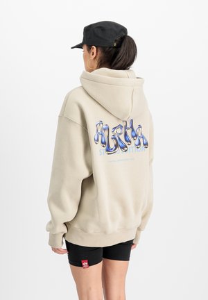 Haut à capuche beige oversize avec un graphisme "ALPHA" en bleu et argent au dos. Assorti à un short noir. Casquette noire portée à l'envers.