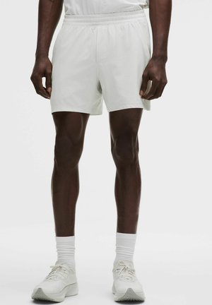 Mannelijk model draagt witte sportshorts, witte sokken en witte hardloopschoenen, staand tegen een effen achtergrond.