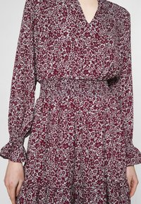 Långärmad bordeauxblommig klänning med V-hals, smockad midja och volangärmar. Har ett ljusgrått blommönster.