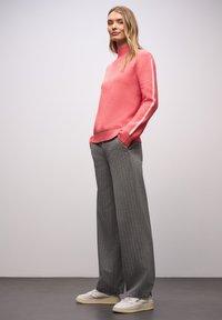 Koralově růžový svetr s vysokým límcem a bílými pruhy na bocích, v kombinaci s šedými širokými kalhotami s proužky a bílými teniskami.