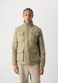 Jachetă khaki cu guler înalt, multiple buzunare frontale și închidere cu fermoar. Fabricată dintr-un material neted, ușor.