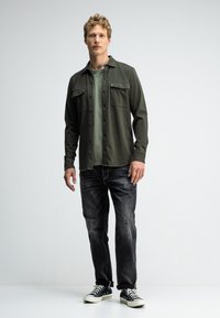 Olivgrünes Hemd mit Knopfleiste und zwei Brusttaschen, über einem hellgrünen T-Shirt getragen, kombiniert mit schwarzen Jeans und schwarzen Sneakers.
