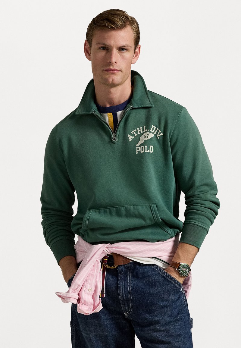 Felpa verde scuro con colletto a mezze zip e logo bianco. Indossata sopra una camicia a righe blu e gialle, abbinata a jeans scuri e un foulard rosa chiaro legato in vita.