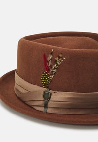 Brun filthat med beigerand och dekorativa flerfärgade fjädrar som hålls av en liten sköldformad metallpin.