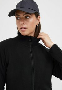 Musthave fleecetagi kõrge kraega ja tõmblukuga. Kandmine tumesinise mütsiga. Sile tekstuur, minimaalne brändimine rinnal.