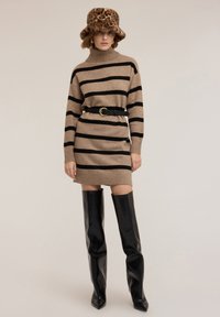 Abito maglione a maglia con righe beige e nere, caratterizzato da un collo alto, cintura nera con fibbia dorata, abbinato a stivali neri al ginocchio.