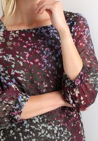 Femme les bras croisés portant un chemisier à manches longues à motifs floraux avec des nuances de violet, bleu et blanc sur un fond sombre.