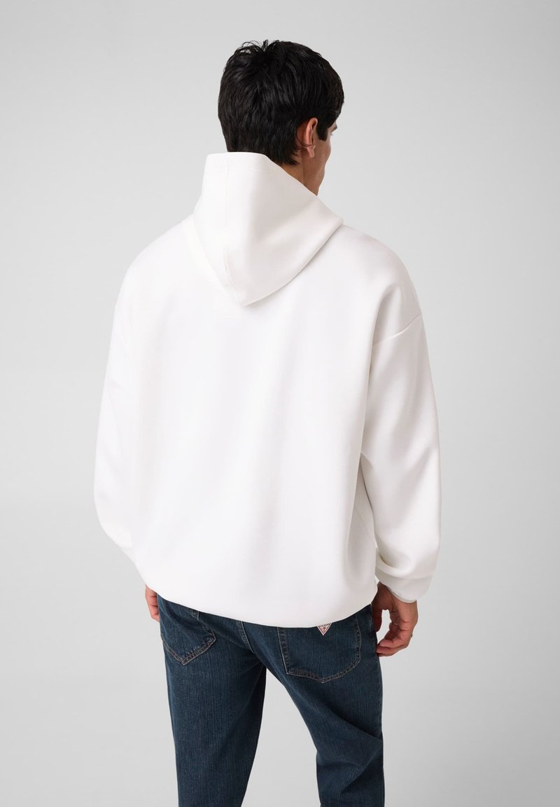 Homme de dos portant un sweat à capuche blanc uni et un jean bleu sur un fond clair uni.