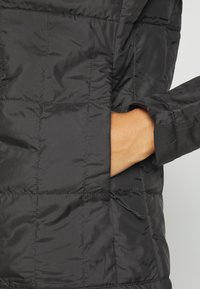 Main partiellement dans la poche latérale d'une veste matelassée noire avec des coutures visibles et un cordon ajustable.