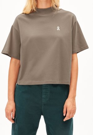 Vrouw draagt een loszittende bruine korte-mouwen top met een klein wit logo op de borst en donkergroene broek.