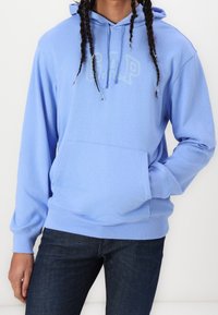 Personne portant un sweat à capuche GAP bleu clair avec une poche avant et un jean bleu foncé, les mains détendues, les cheveux tressés, se tenant contre un fond uni.