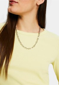 Femme portant un t-shirt jaune côtelé et un collier en chaîne argenté avec des maillons alternés circulaires et allongés.