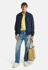 Marine bomberjakke over en gul polo, blå jeans og hvide sneakers. Holder en lys totebag. Casual, lagdelt outfit med kontrasterende farver.