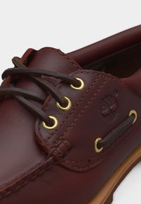 Chaussure bateau en cuir marron avec des lacets marron foncé et des œillets dorés, présentant des détails cousus et un logo Timberland embossé sur le côté.