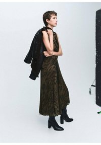 Vestido marrón plisado con un patrón abstracto oscuro, combinado con una chaqueta negra caída sobre el hombro y botas de tacón negras.