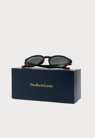Fekete ovális napszemüveg szürke lencsékkel, texturált fekete gumi kerettel, piros részletekkel, egy sötétkék dobozra helyezve, amelyen arany "Polo Ralph Lauren" logó található.
