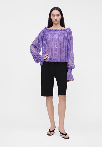 Femme portant un chemisier en dentelle violette transparente à épaules dénudées, un short noir longueur genoux et des sandales noires à talons, debout avec une main sur la hanche.