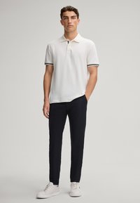 Polo blanc avec des manches rayées, associé à un pantalon sombre et des baskets blanches. La chemise a un tissu texturé et une coupe détendue.
