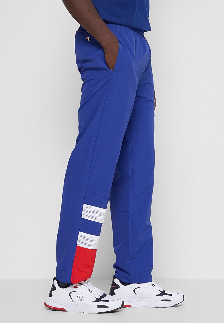 Pantalons de sport bleus avec une taille élastique, poches latérales, présentant des rayures en maille blanche et un accent rouge. Portés avec des baskets blanches.