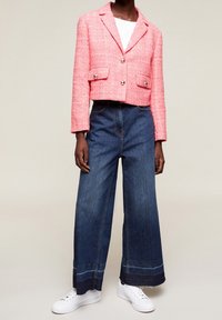Różowa krótka marynarka z tkaniny tweedowej z złotymi guzikami, zestawiona z wysokim stanem, szerokimi nogawkami ciemnoniebieskich dżinsów oraz białymi tenisówkami.