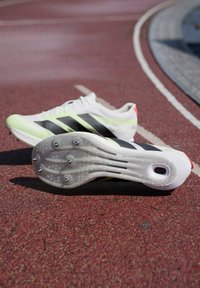Chaussures de sport légères blanches avec des rayures noires, des accents verts, et une semelle extérieure texturée munie de six pointes métalliques pour une meilleure adhérence.
