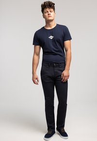 Granatowy t-shirt z białym logo, w połączeniu z ciemnymi dżinsami. Model ma na sobie niebieskie buty z pomarańczowymi sznurowadłami, stoi na tle jednolitego tła.