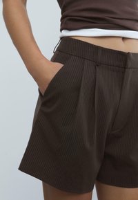 Shorts marrones con rayas de pespunte que presentan un diseño a medida, pliegues y bolsillos laterales inclinados. Fabricados con una textura de tela suave.