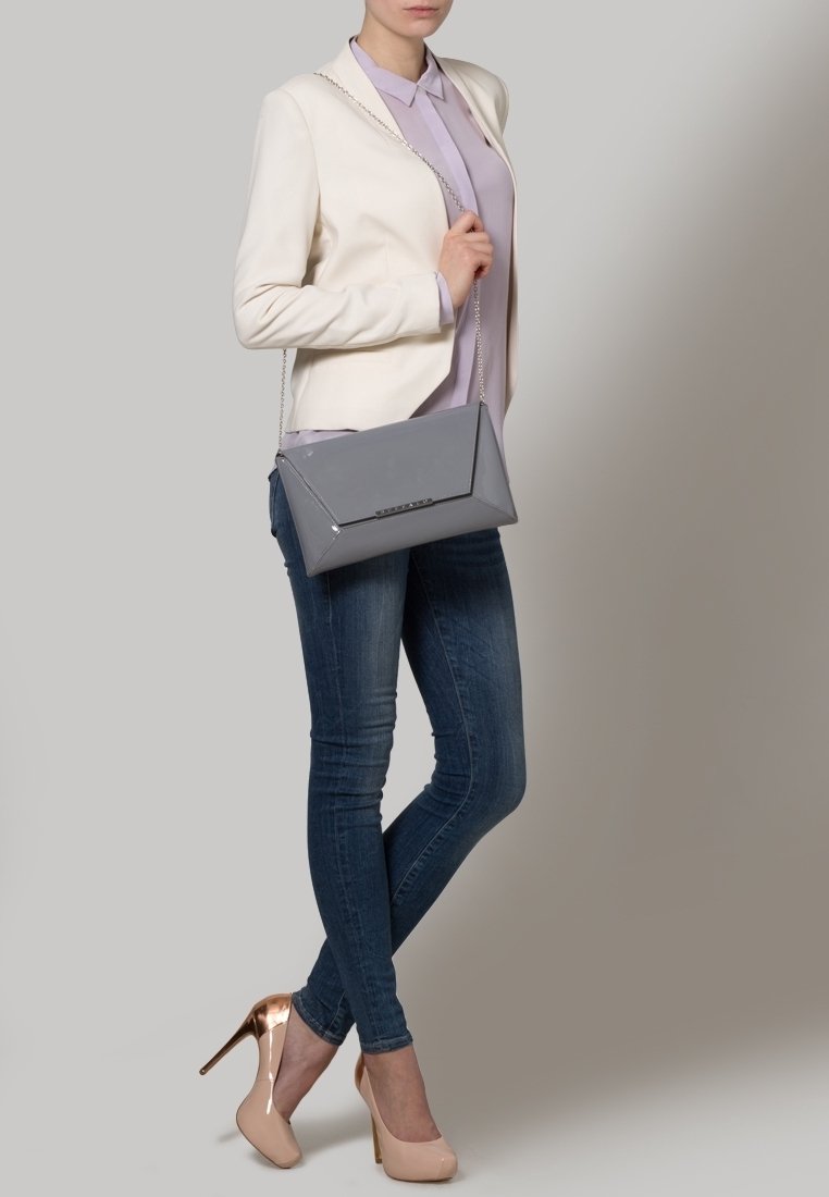 Graue lackierte Clutch mit geometrischem Design, Kettenriemen und einem Klappenverschluss. Kombiniert mit einer creamfarbenen Jacke und einem helllila Hemd.