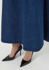 Jupe en denim bleu foncé avec un design long et fluide, avec des détails de couture visibles à l'ourlet. Assortie à des escarpins noirs à talons aiguilles.