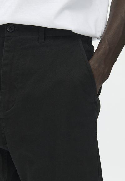 Pantalon en coton noir à coupe droite, doté de poches latérales et d'une fermeture par bouton. Le tissu présente une texture lisse et des détails de couture minimes.