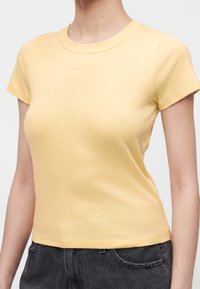Personne portant un t-shirt ajusté jaune pâle à manches courtes et un jean foncé, montrée du cou à la taille sur un fond uni.