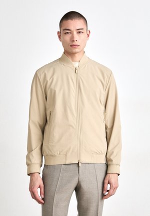 Beige bomberjakke med frontglidelås, elastiske mansjetter og stikklommer. Laget av glatt stoff, med standkrage og minimalistisk design.