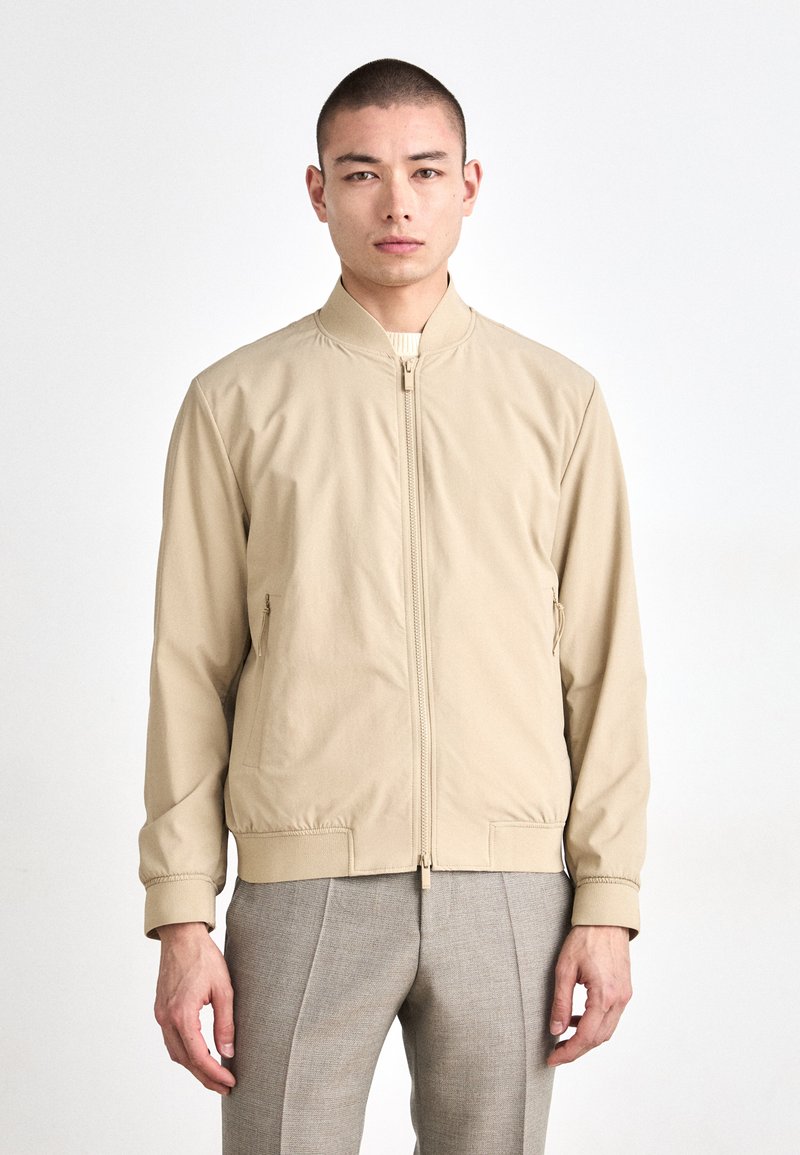 Beige bomberjakke med frontglidelås, elastiske mansjetter og stikklommer. Laget av glatt stoff, med standkrage og minimalistisk design.
