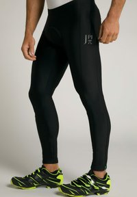 Schwarze Sportleggings mit einer figurschmeichelnden Passform, die leicht glänzen, mit einem Logo am linken Oberschenkel und Reißverschlüssen an den Knöcheln. Kombiniert mit sportlichen Schwarz- und neongrünen Schuhen.