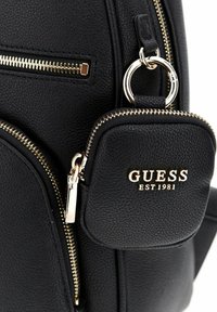 Sac à dos noir texturé avec des détails en or, comprenant une petite pochette zippée attachée, estampillée avec "GUESS EST 1981" en lettres dorées.