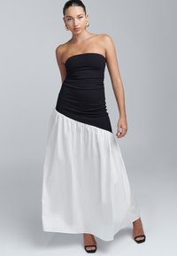 Melna strapless kleita ar pieguļošu augšdaļu, kas pāriet plūstošā baltā maksi svārkā, ar teksturētu viļņotu apakšmalu un elegantu siluetu.