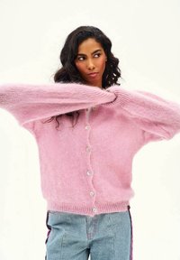 Žena s dlhými tmavými vlasmi, oblečená v ružovom nadýchanom kardigáne na gombíky a svetlomodrých džínsach, si upravuje rukáv na jednoduchom pozadí.