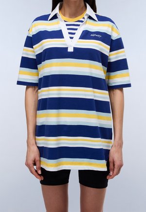 Persoon draagt een loszittend kortarmig shirt met brede horizontale strepen in blauw, geel, wit en lichtblauw, en een zwarte korte broek.