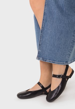 Piernas con falda vaquera azul y zapatos planos Mary Jane de cuero negro con correas en el tobillo y hebillas plateadas sobre fondo blanco.