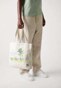 Lerretstrålepose med trykt strandmotiv som viser et palmetre, sol og strand i lys beige. Person iført lysegrønn topp og beige bukser.