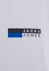 Bijela pamučna majica s printanim "JACK&JONES" logotipom. Logotip sadrži horizontalni plavi naglasak i crne detalje.