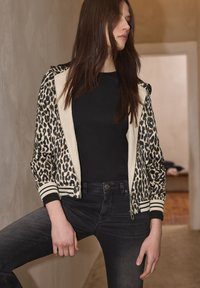 Bomberjacke mit Leopardenmuster, cremefarbenem Vorderteil, gerippten Bündchen und Saum. Getragen über einem schwarzen Oberteil mit dunklen Jeans. Glatte Stoffstruktur.